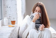 young woman with flu looking at thermometer and blowing nose in tissue